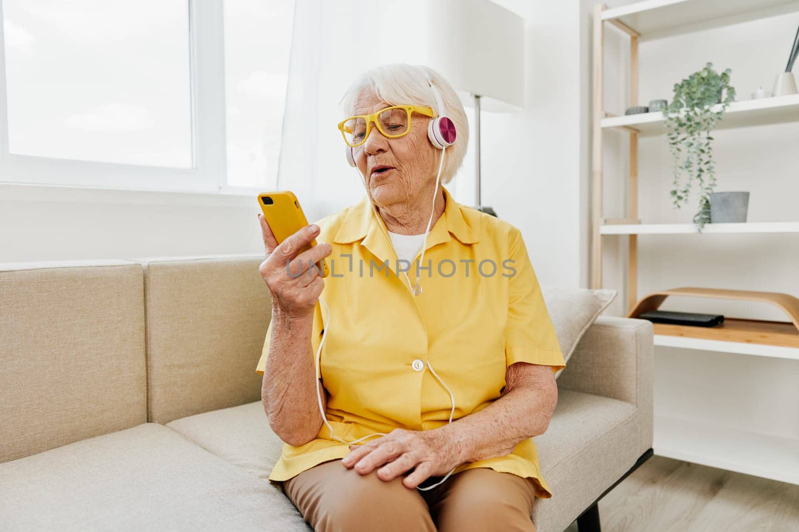 Happy elderly woman looking into her phone video call with headphones smile, technology for communication, bright modern interior, lifestyle online communication. High quality photo