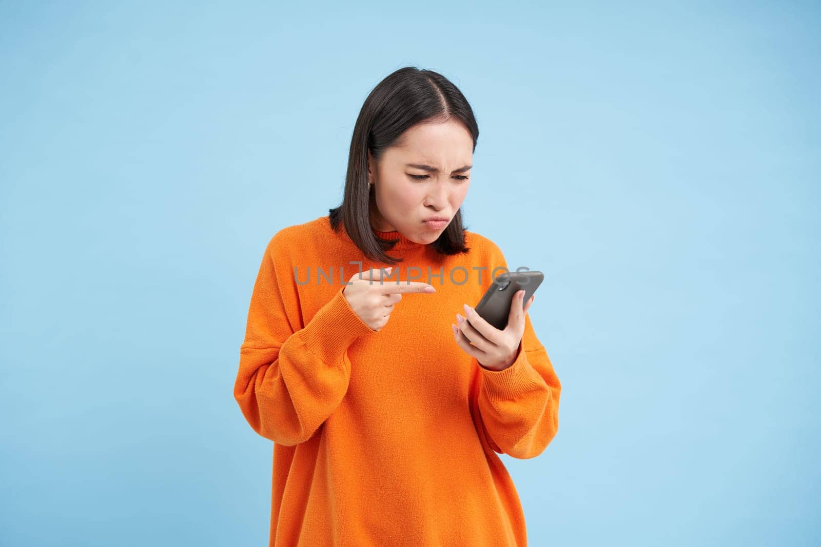 Annoyed girl with phone, points at mobile screen with frowning angry face, standing disappointed over blue background.