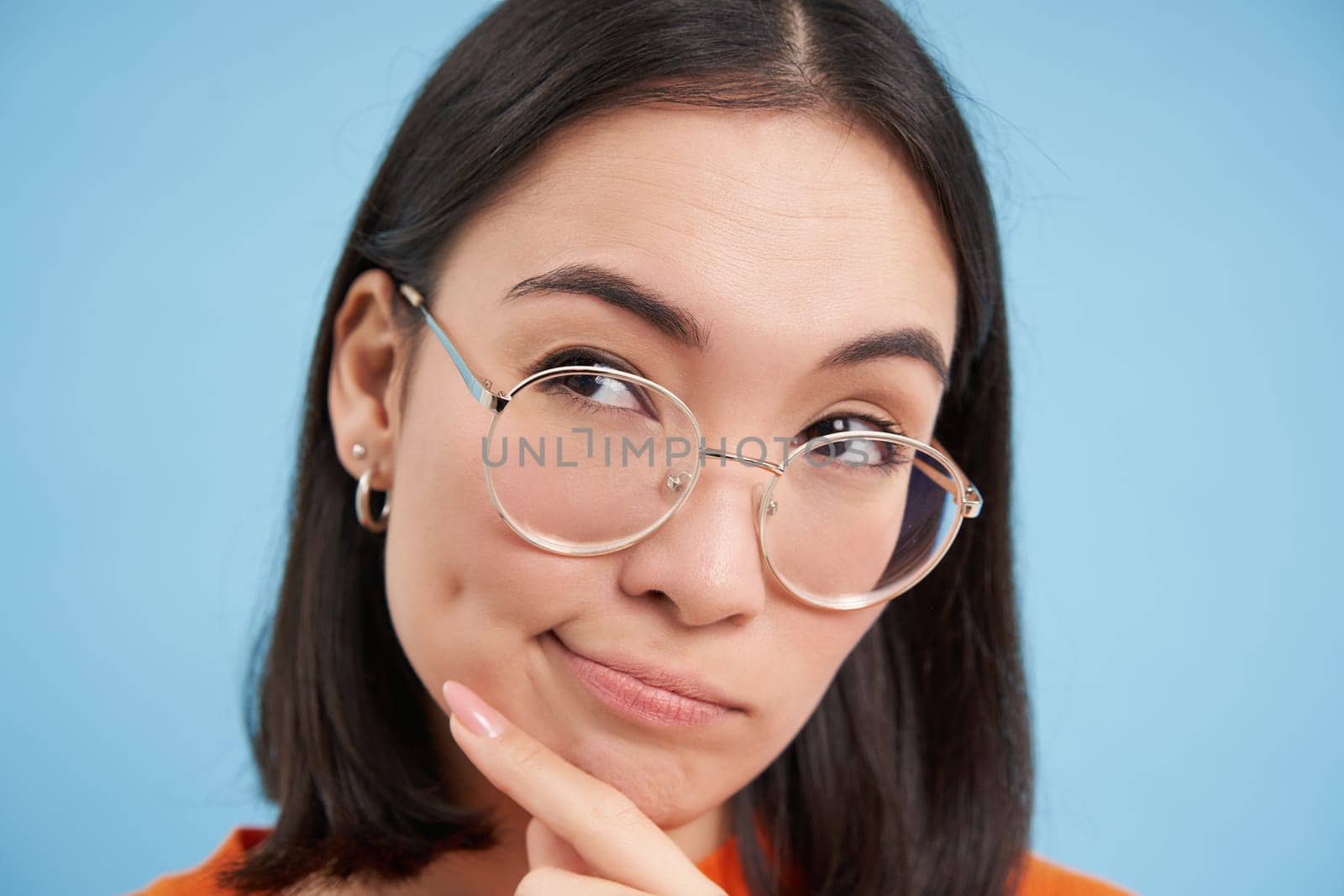 Portrait of Japanese woman in glasses, looks thoughtful, ponders, thinking with serious face, standing over blue background.