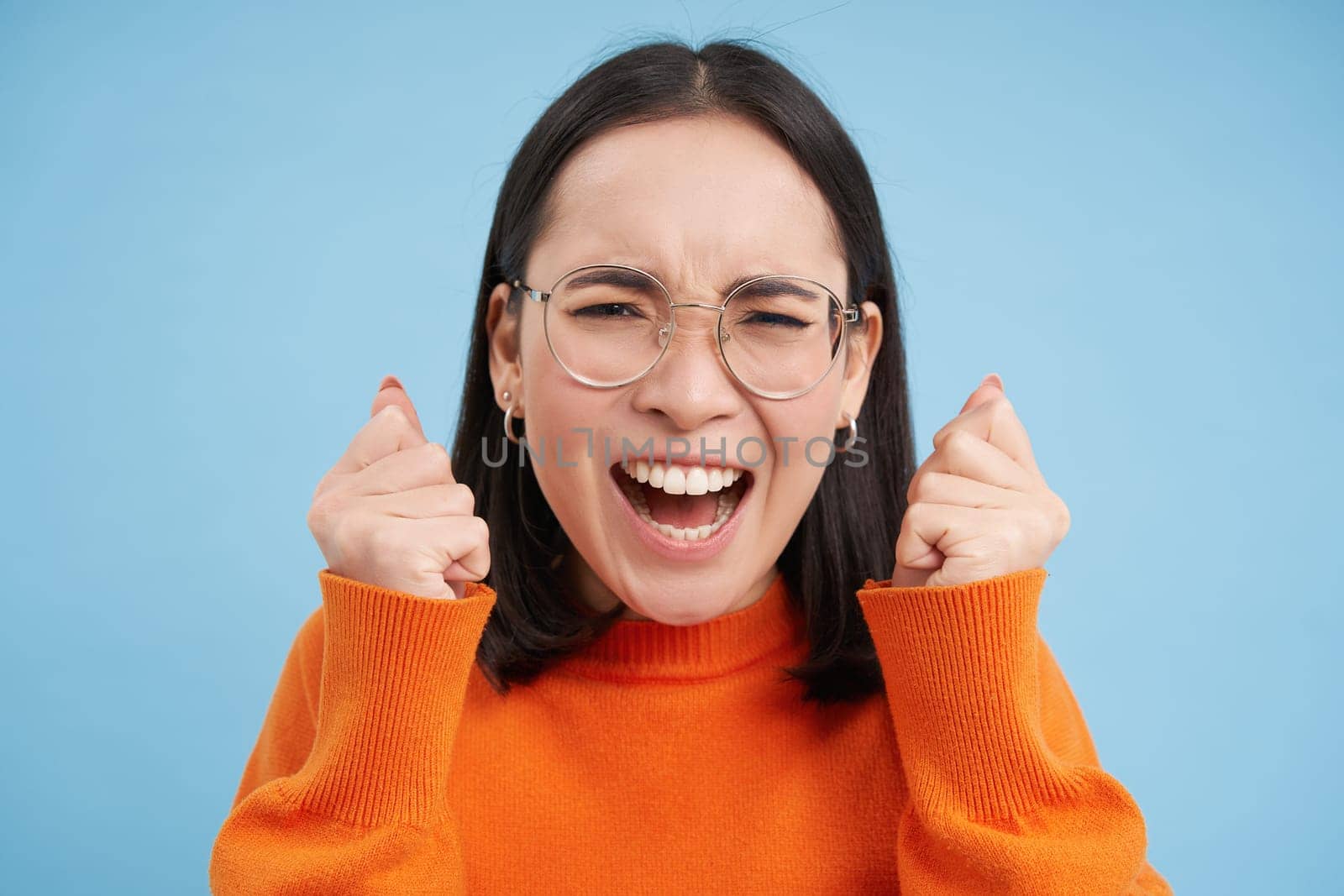 Portrait of young asian woman in glasses, arguing, screaming and looking annoyed, standing over blue background.