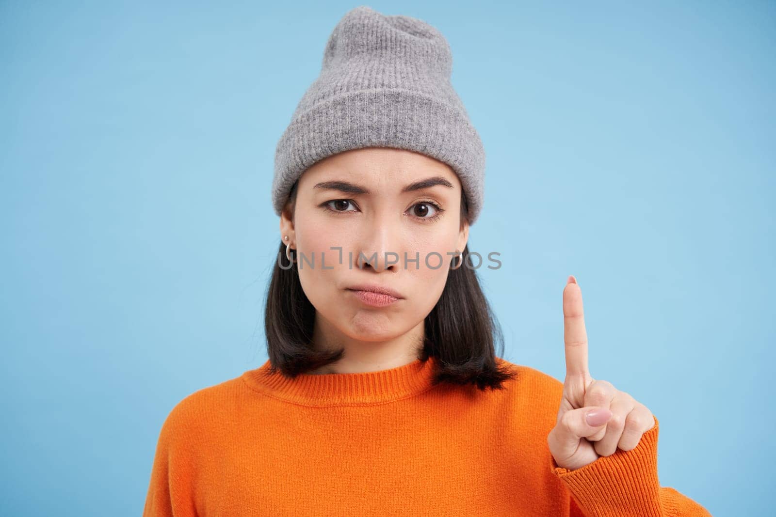 Asian girl in hat scolding you, shakes finger in disapproval, disappointed by behavious, standing over blue background.