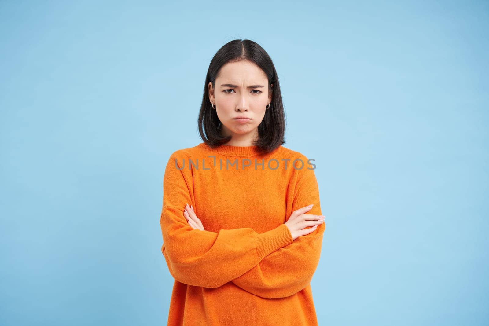 Defensive asian woman shuts herself from conversation, cross hands on chest, sulks and frowns with angry face, stands over blue background