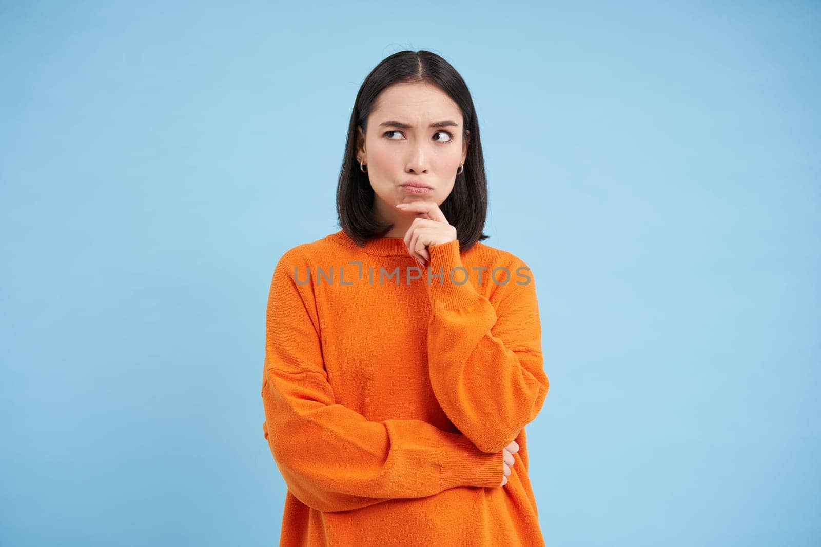 Brainstorm, ideas concept. Portrait of asian girl thinks and looks puzzled, stands thoughtful in orange sweatshirt, makes decision, blue background by Benzoix