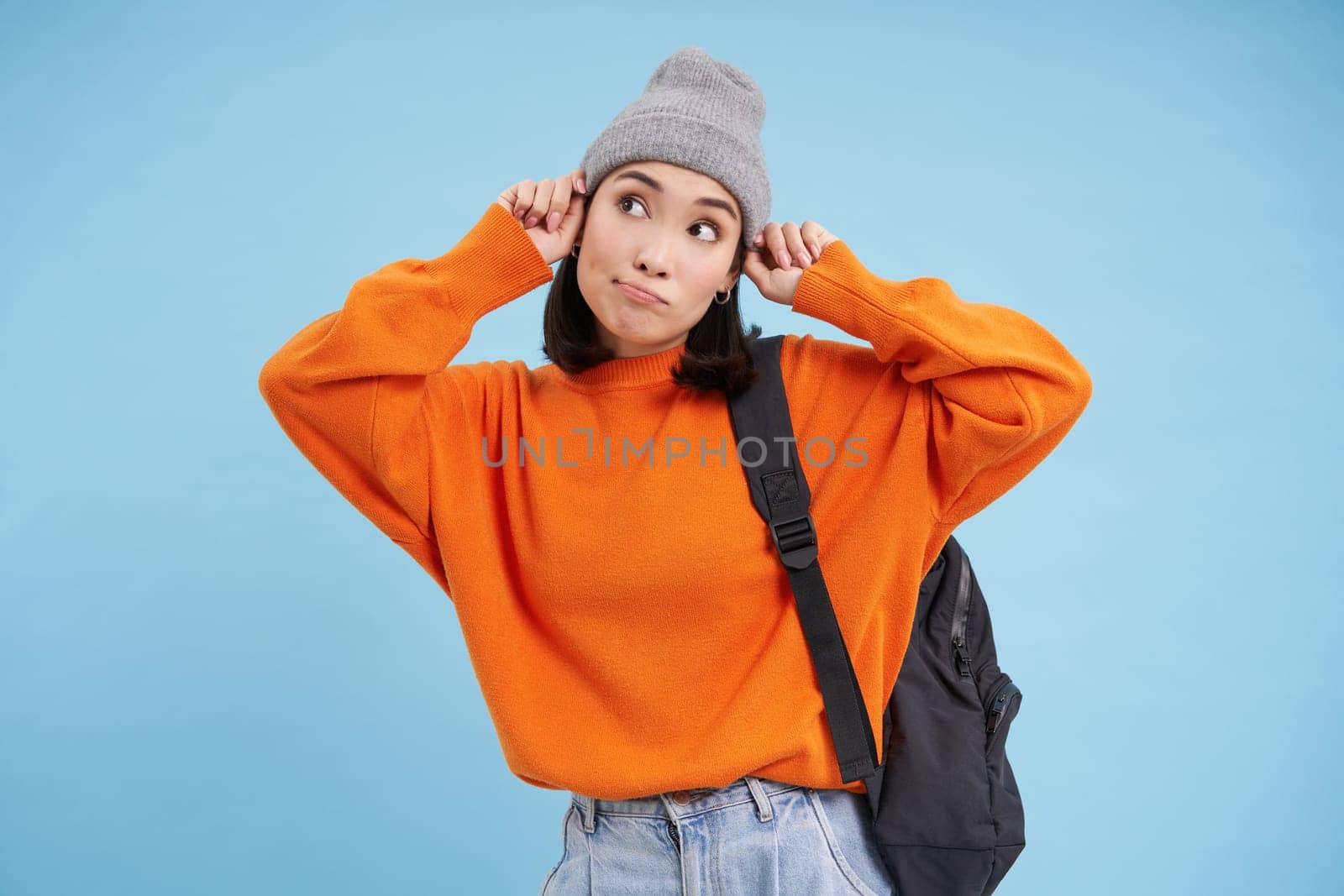 Happy stylish korean woman puts on hat, goes for walk with backpack, stands over blue studio background.