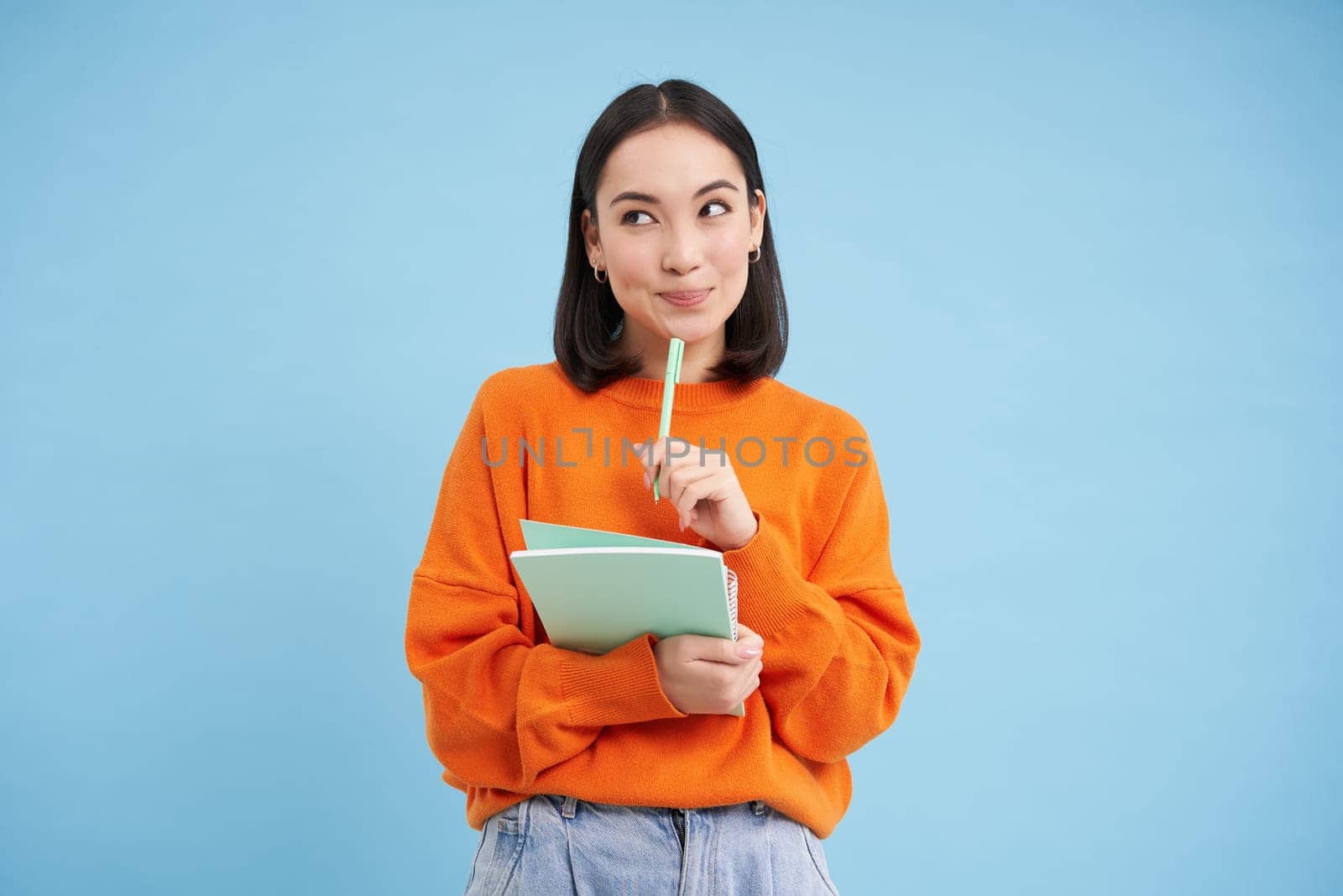 Education and students. Happy asian woman, holding notebooks and laughing, smiling at camera, enjoys going to University or College, blue background by Benzoix