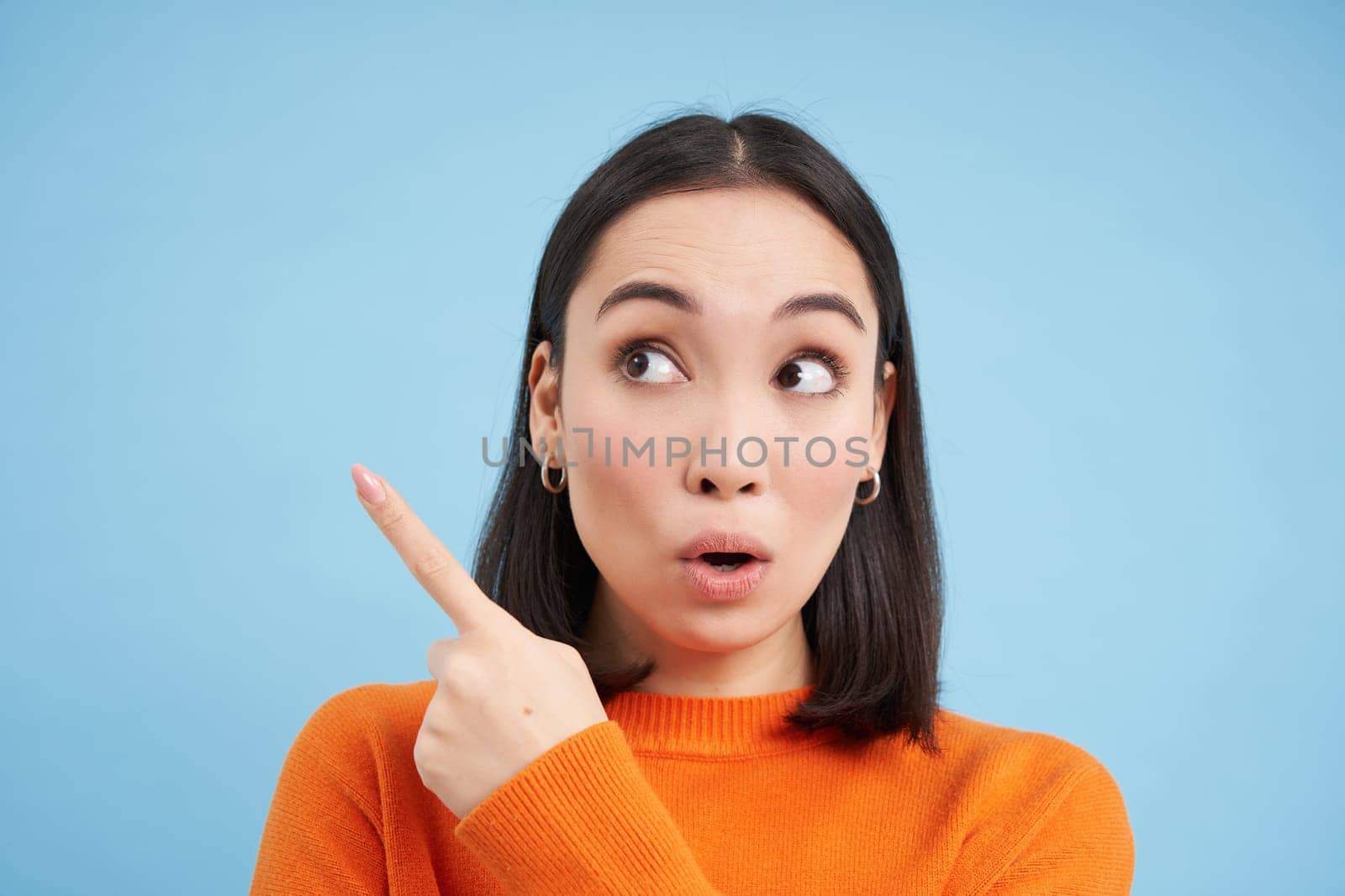 Close up portrait of asian girl, looks surprised, points at smth interesting, amazed by advertisement, blue studio background.