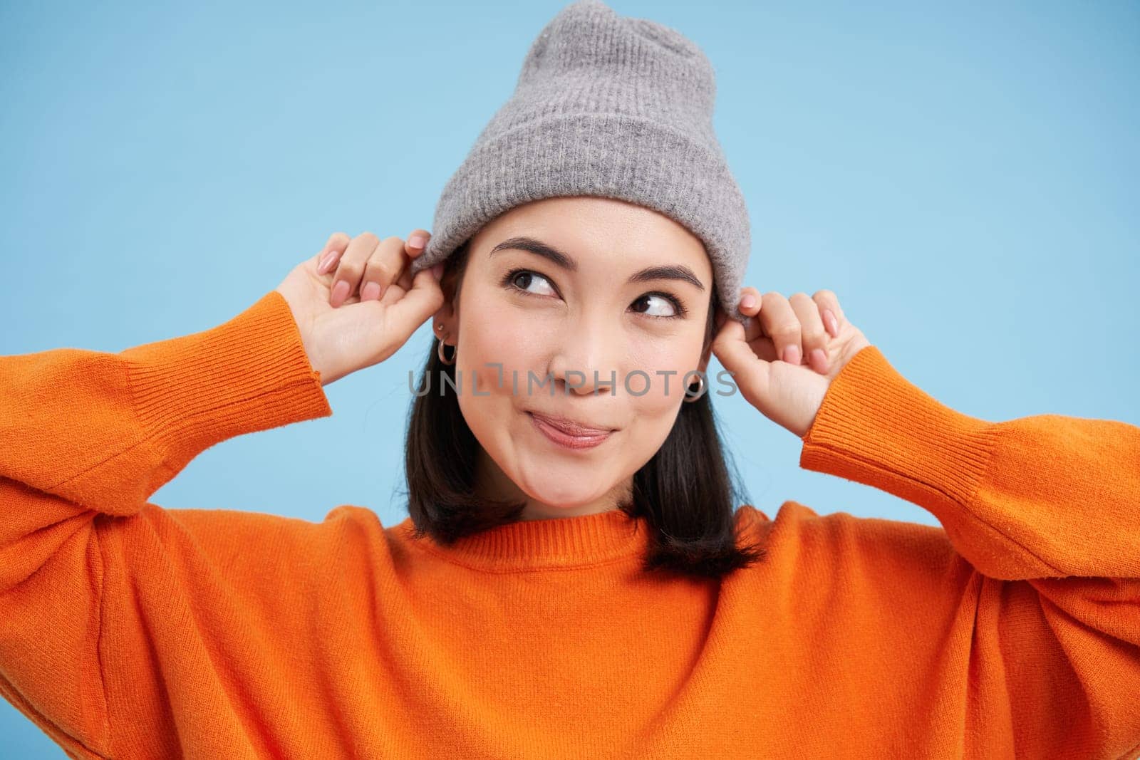 Close up portrait of happy, stylish korean girl puts on warm hat, smiles and looks joyful, stands against blue studio background.