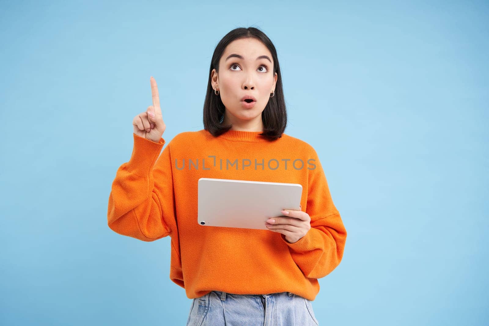 Girl with surprised face, pointing and looking up, holding digital tablet, showing awesome promo offer, standing over blue background.