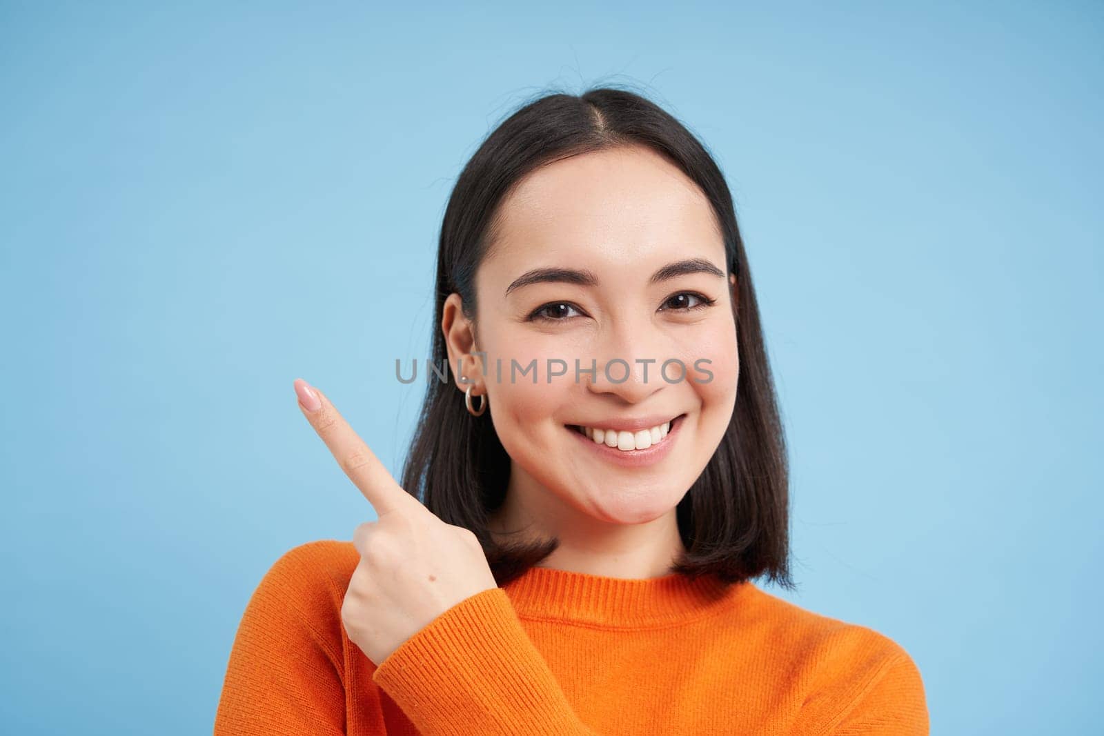 Close up of lovely girl points finger at banner, shows copy space, stands over blue background by Benzoix