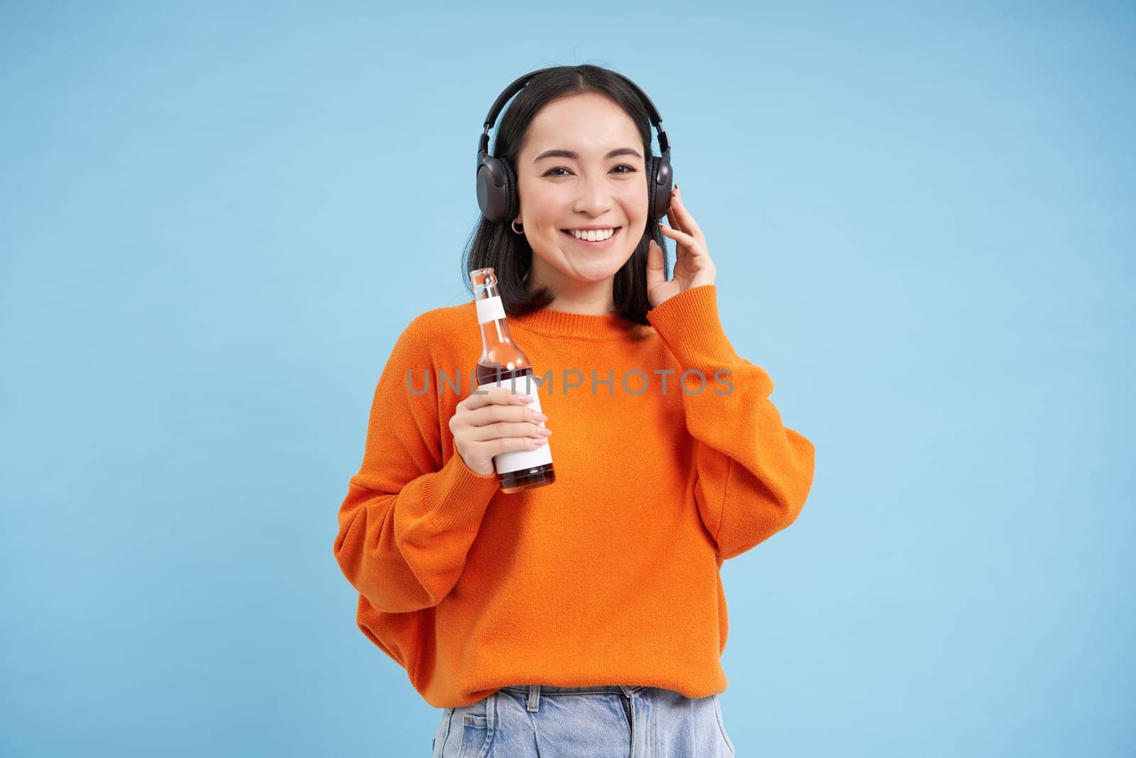 Beautiful asian woman listens music in headphones, drinks coke from bottle and smiles at camera, blue background.