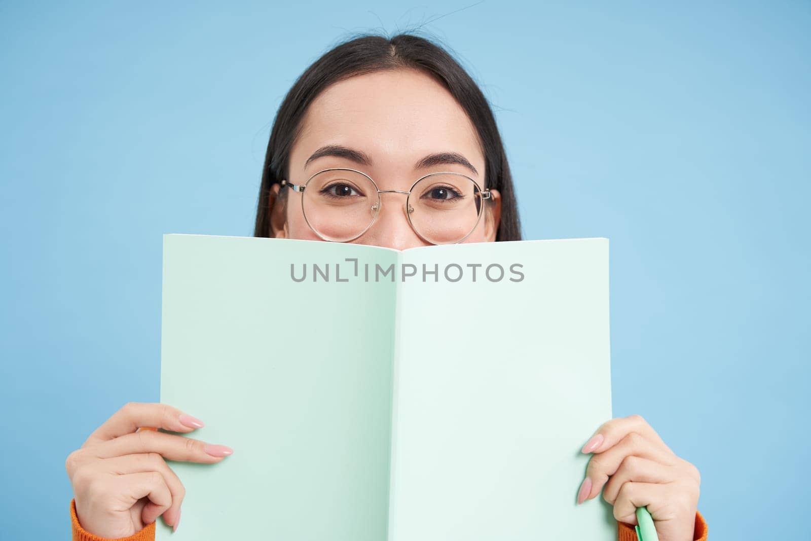 Portrait of cute japanese woman in glasses, peeking from notebook, covers face with her notes, smiles with eyes, blue background by Benzoix