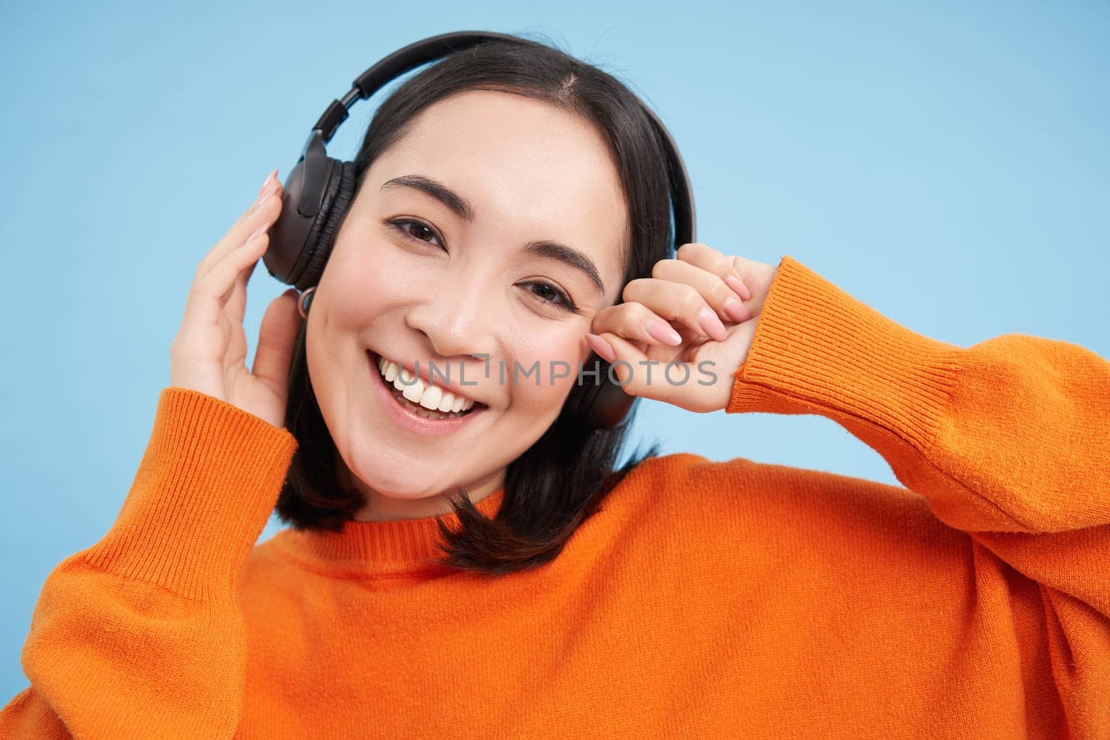 Close up of beautiful asian woman in headphones, smiles and listens music, enjoys podcast, stands over blue background.