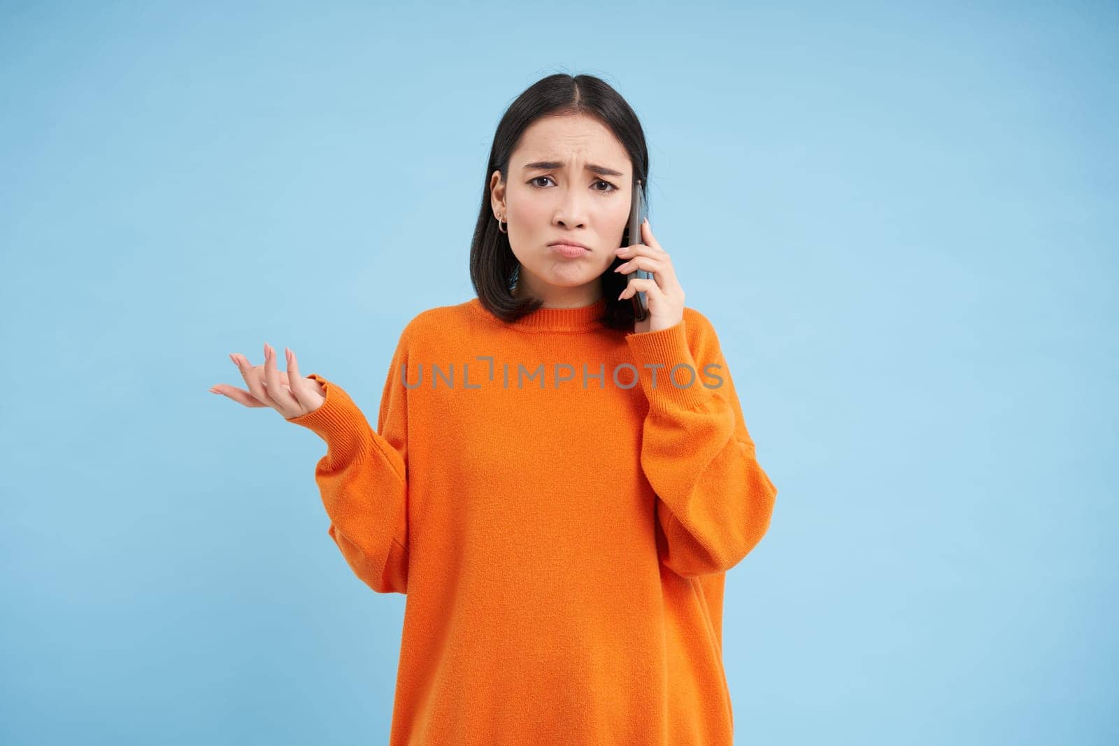 Sad and confused asia girl shrugs, talks on cellphone, answers telephone call with puzzled face, stands over blue background.