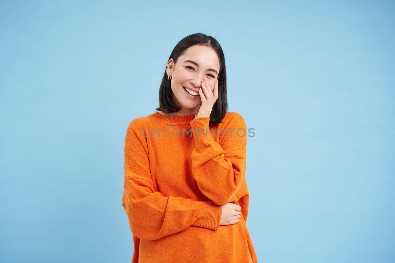 Beauty and wellbeing. Young brunette woman laughing and smiling, touching her natural healthy face, standing over blue studio background.