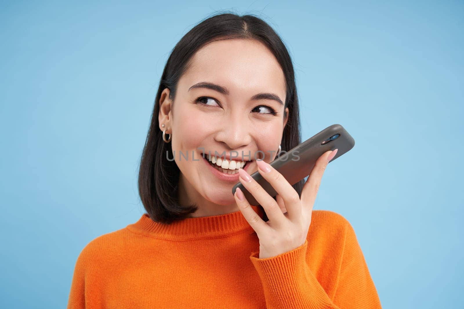 Portrait of asian woman records voice message, talks on speakerphone, holds mobile phone near mouth while speaking, translates her speech on app, blue background by Benzoix