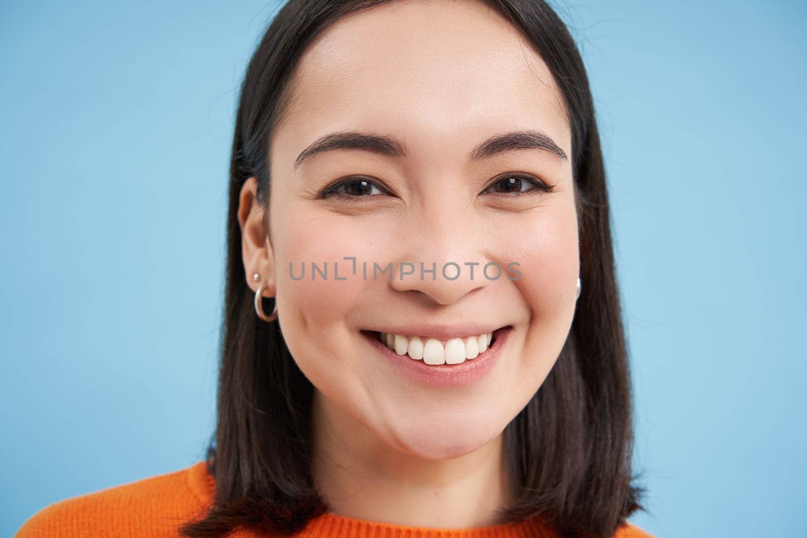 Beauty and wellbeing. Close up portrait of young happy asian woman, smiling and showing candid emotions, standing over blue background.