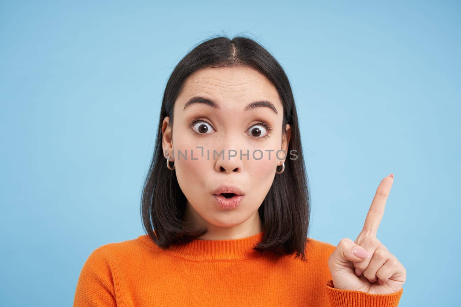 Close up portrait of asian girl, looks surprised, points at smth interesting, amazed by advertisement, blue studio background.