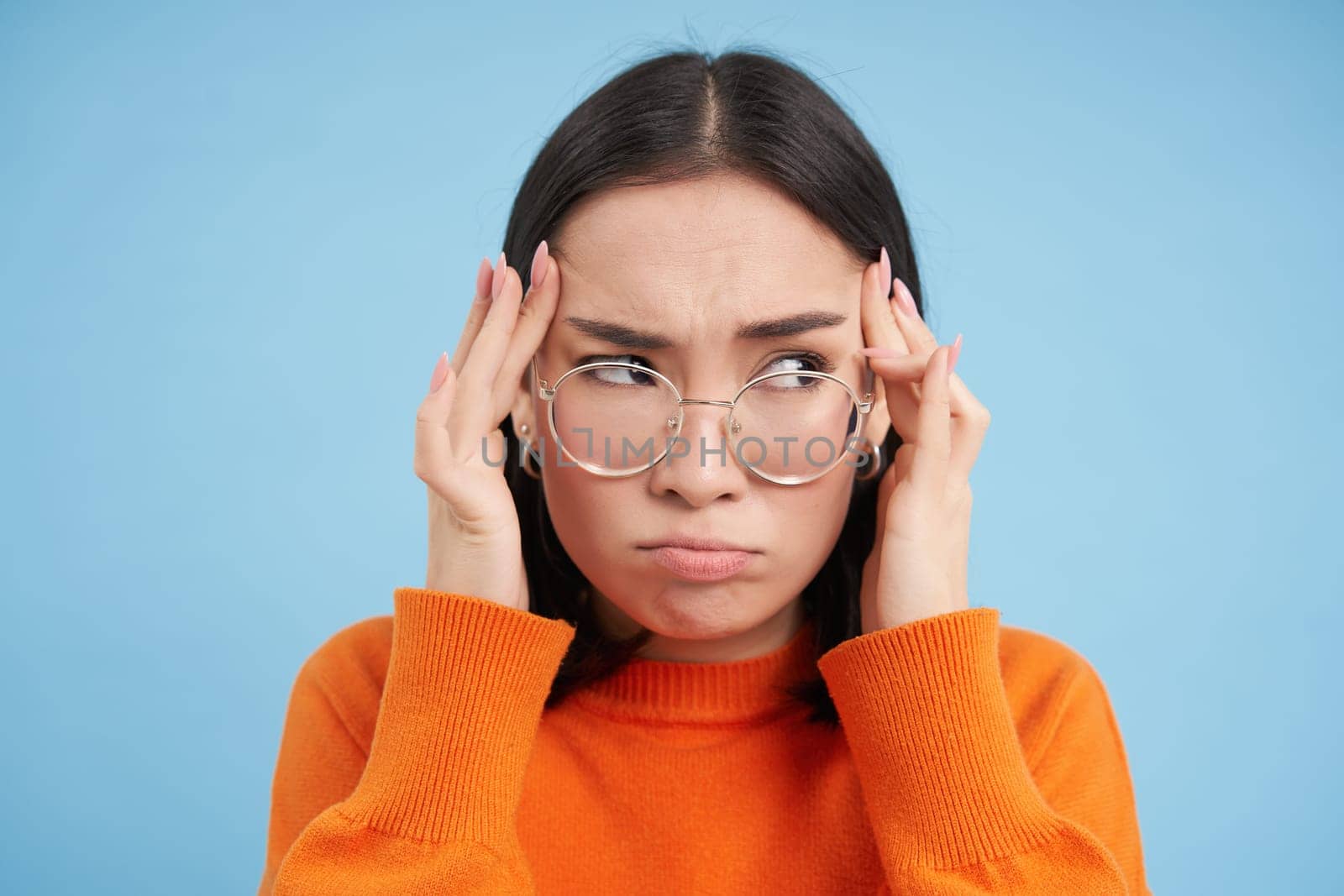 Portrait of asian woman in glasses, holds hands on head complicated face, feels troubled, has problems, standing confused over blue background by Benzoix