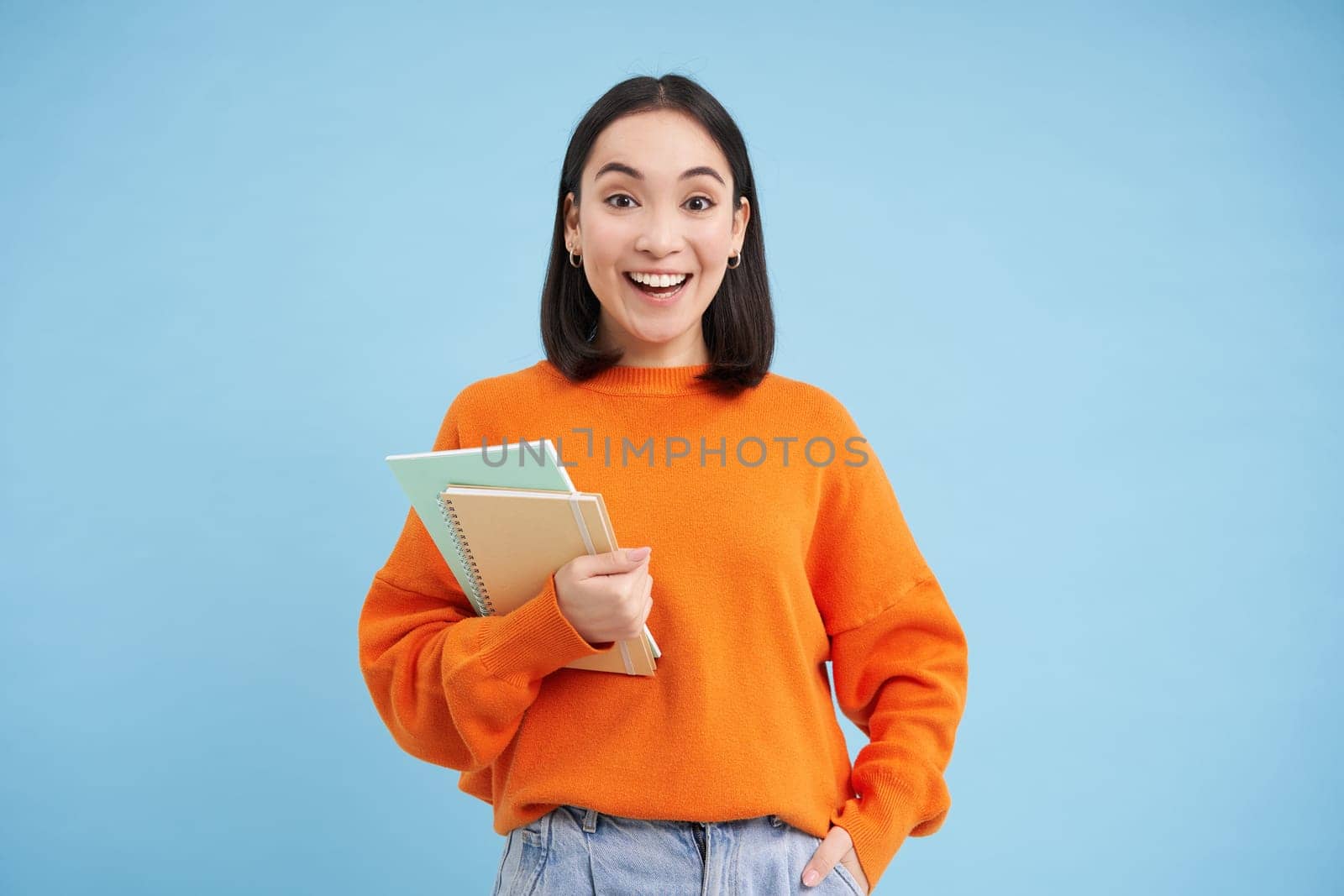 Young asian student with notebooks, standing in orange sweatshirt, looking at camera, posing over blue background by Benzoix