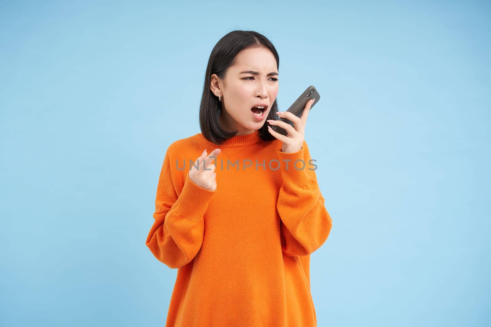 Angry asian woman yells at mobile phone, shouts in smartphone with furious face, stands over blue background by Benzoix