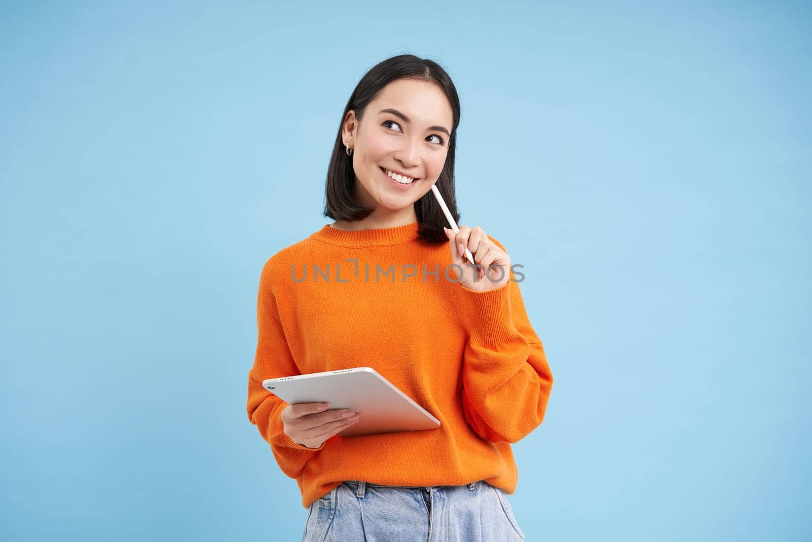 Modern beautiful asian woman with digital tablet and pencil, taking notes, writing on her gadget, doing homework, working, standing over blue background by Benzoix