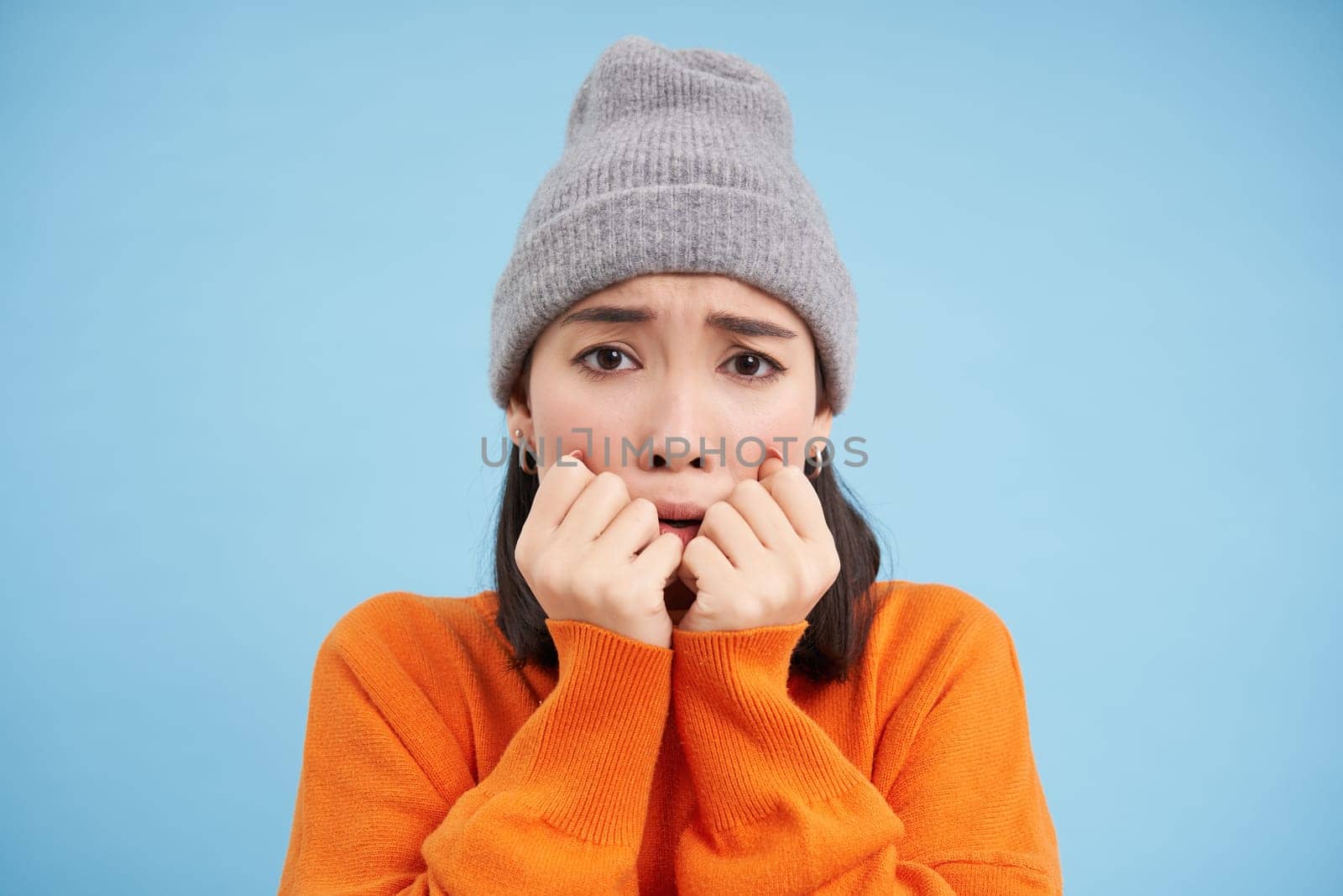 Portrait of asian girl in warm hat, looks with fear, shaking and trembling scared, standing frightened against blue background.