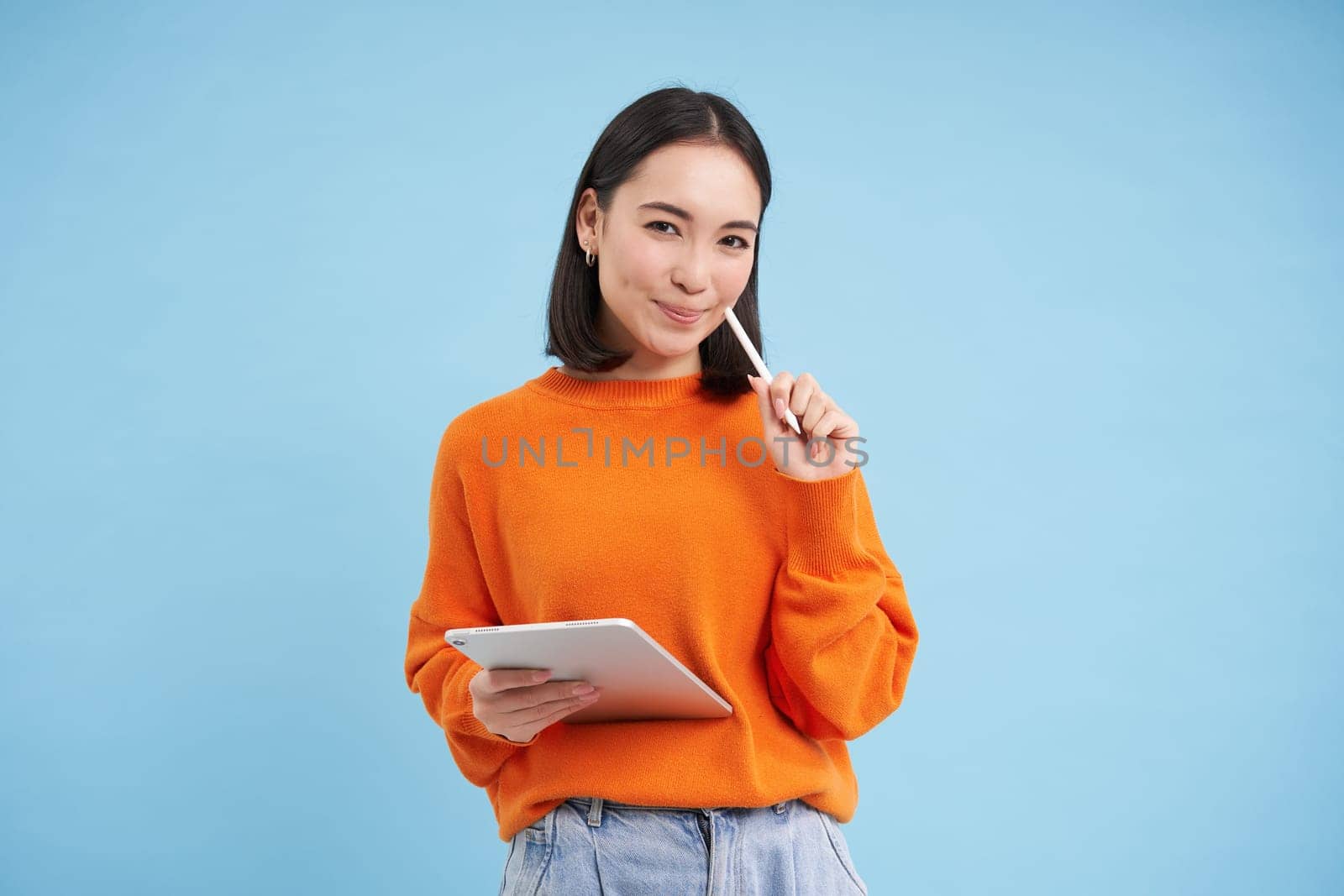 Modern beautiful asian woman with digital tablet and pencil, taking notes, writing on her gadget, doing homework, working, standing over blue background by Benzoix