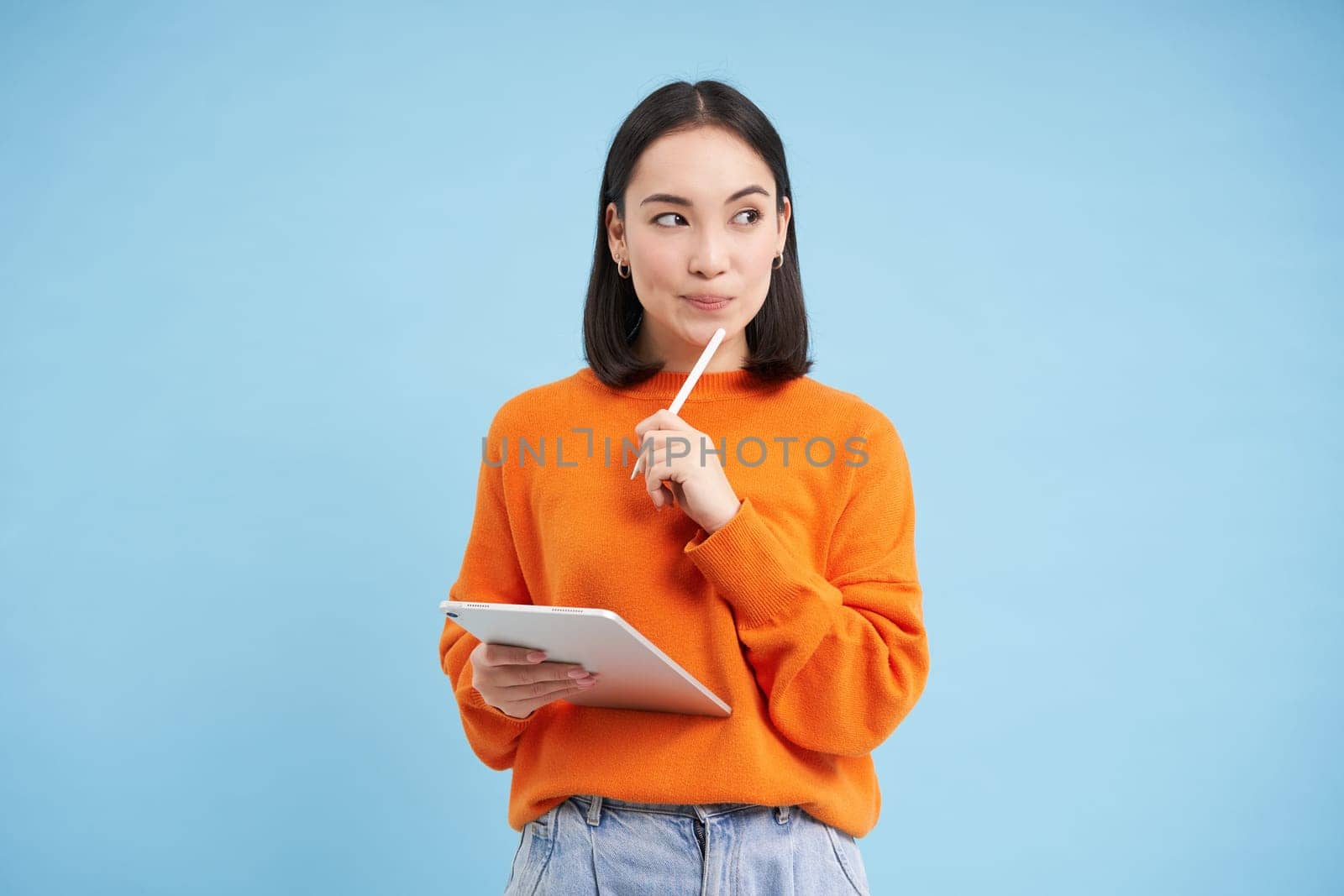 Modern beautiful asian woman with digital tablet and pencil, taking notes, writing on her gadget, doing homework, working, standing over blue background.