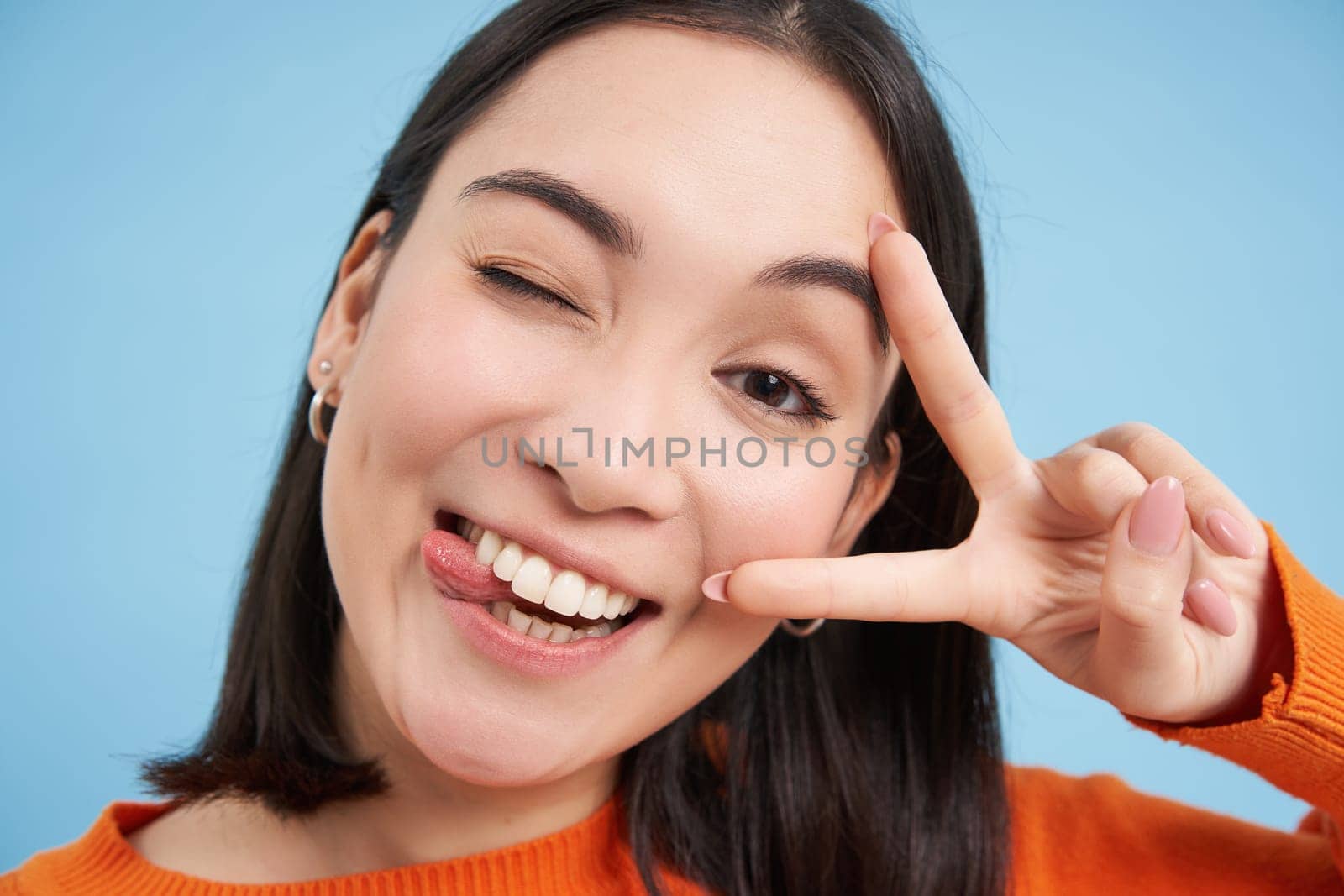 Close up portrait of positive asian woman, winks and shows peave, v-sign gesture, sticks tongue and smiles, stands over blue background.
