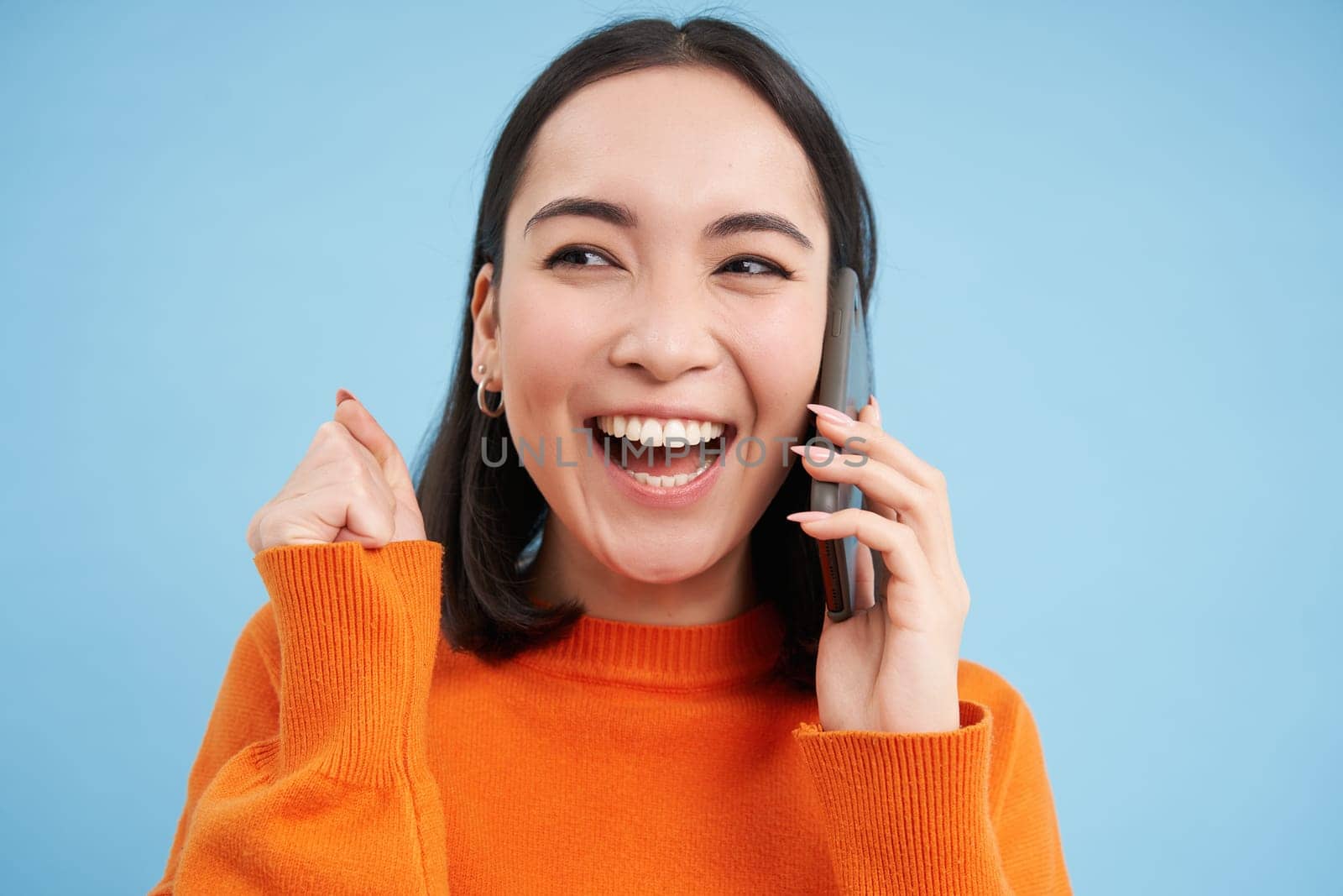 Cheerful asian girl talks on mobile phone, speaks with someone on telephone, stands over blue background. Technology concept