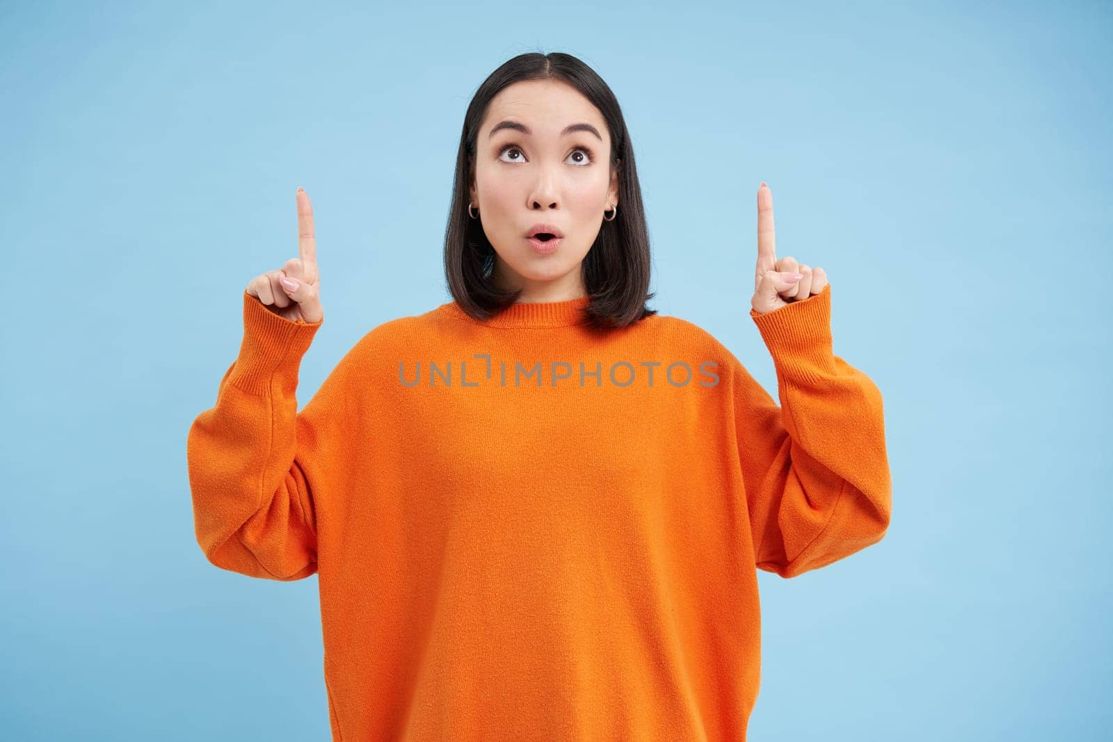 Amazed asian female model, says wow, looks impressed, points fingers up, stands over blue background. Copy space