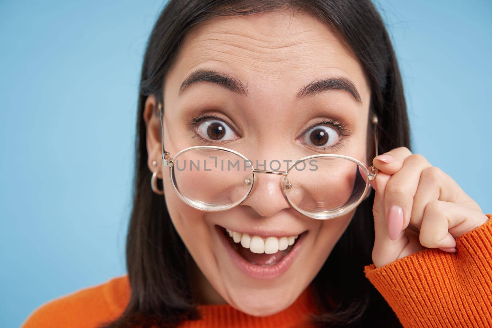 Close up portrait of amazed girl, looks closer at camera in glasses, standing against blue background. Copy space