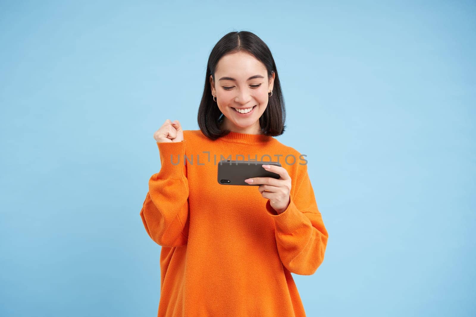 Happy korean girl winning on mobile phone video game, holding cellphone and celebrating, achieve goal in app, standing over blue background.