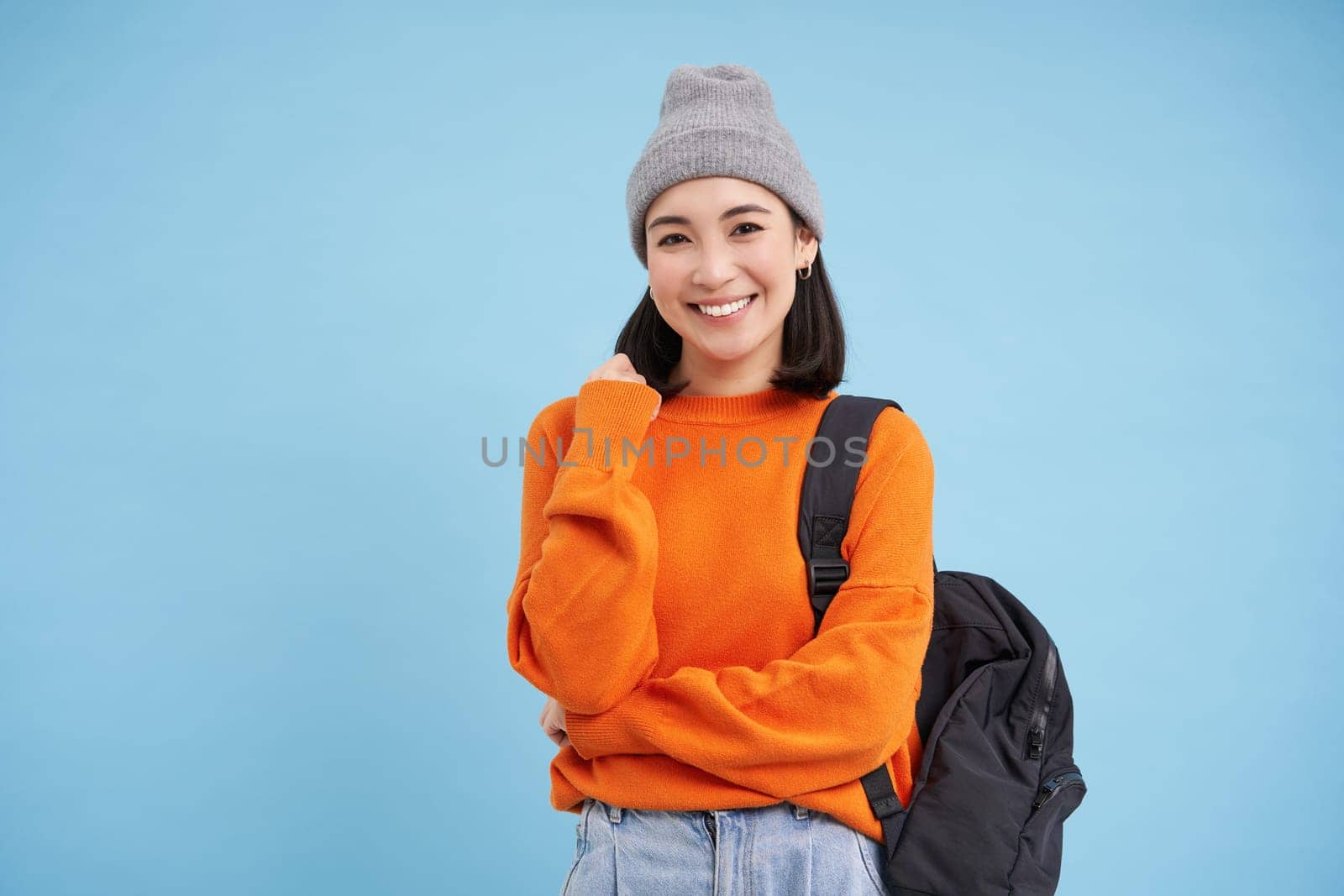 Stylish young asian woman in warm hat, walking with backpack, going somewhere with bag, standing over blue studio background.