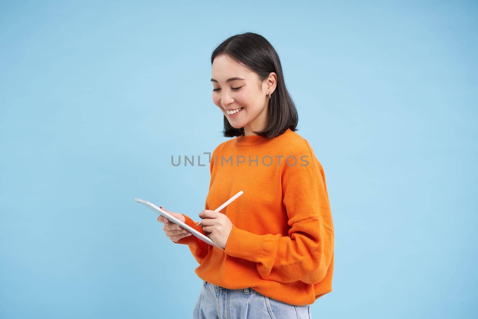 Beautiful modern chinese woman with digital tablet and pencil, looking at screen, writing down notes, doing homework, drawing on gadget, blue background.