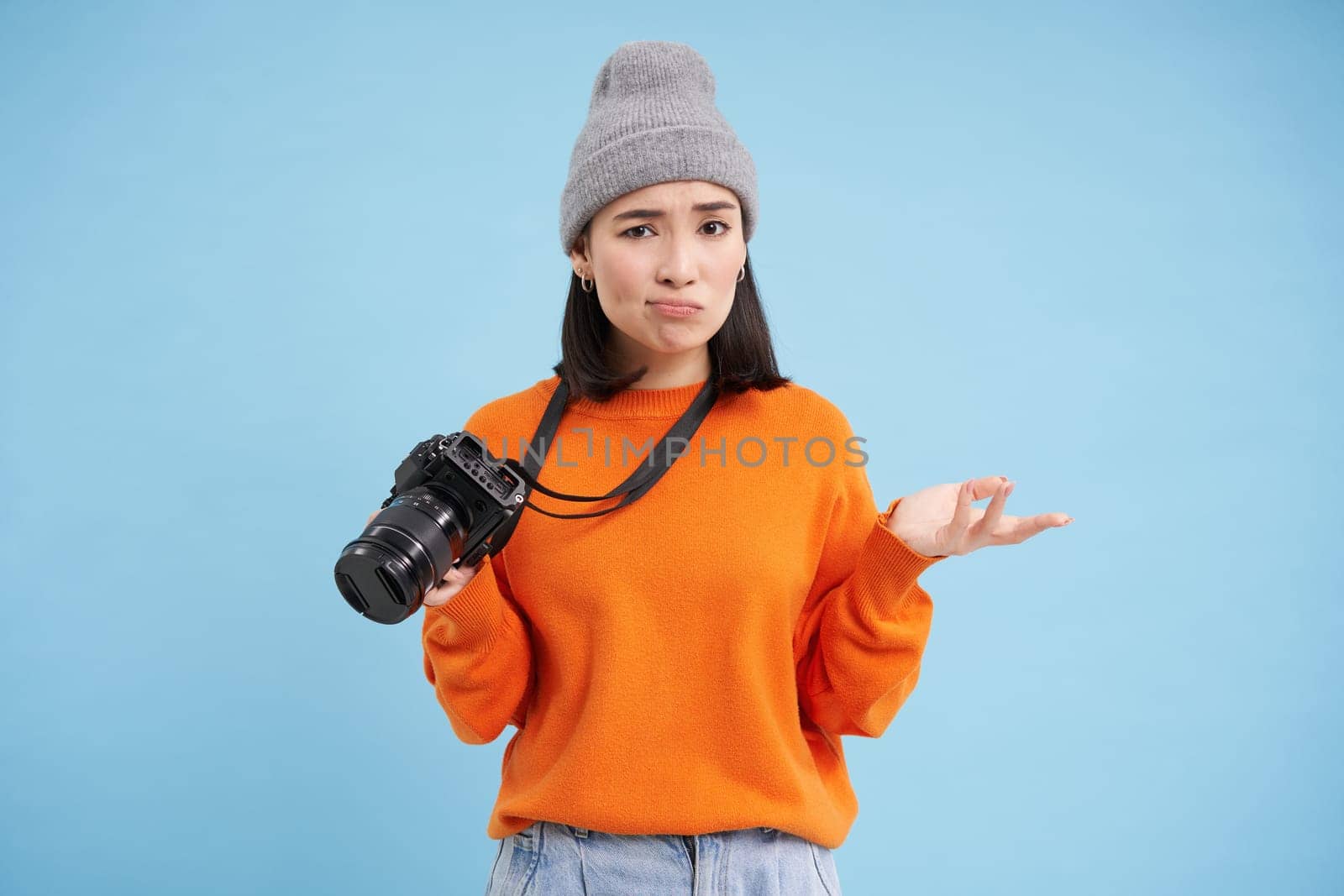 Portrait of asian woman in hat, holding digital camera with confused face, unprofessional photographer doesnt know how to take pictures on digicam, blue background by Benzoix