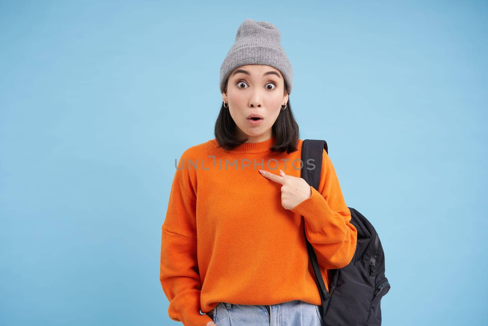 Surprised asian woman points at herself with amazed face expression, looks impressed, stands over blue background by Benzoix