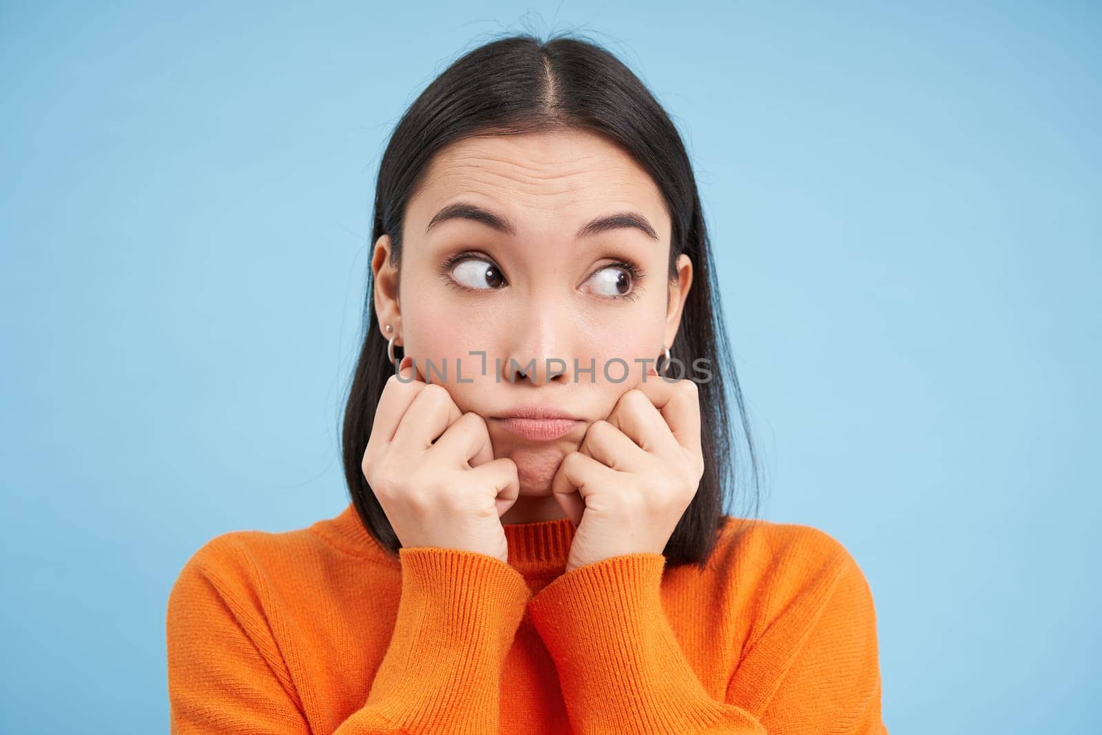 Close up of cute korean girl, looks aside and poses silly against blue studio background. Copy space