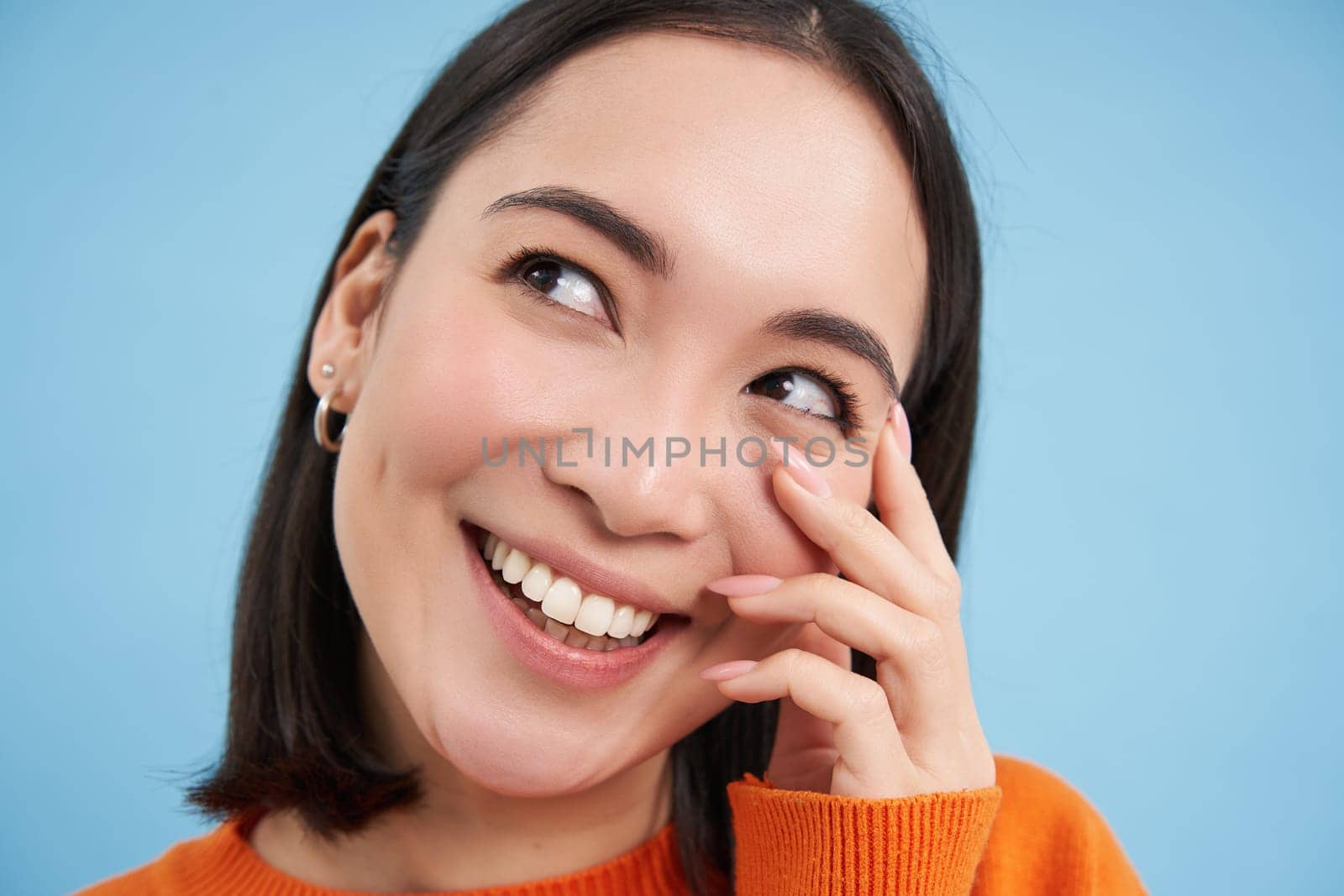 Close up portrait of beautiful japanese woman, smiling and laughing, touches her face, standing over blue studio background.