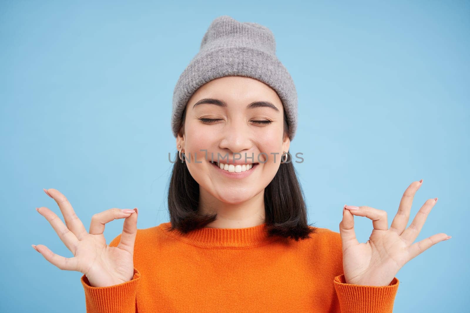 Sounds mind in healthy body. Smiling calm and relaxed asian girl in beanie, shows zen, relaxation gesture, meditating, standing over blue background.