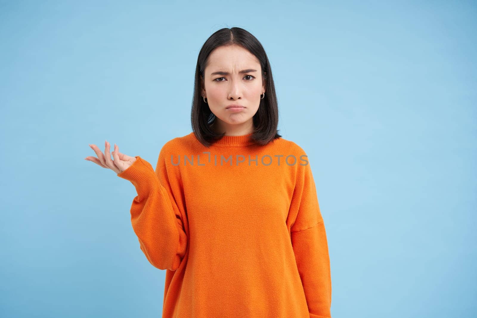 Puzzled asian woman shrugs, raises one hand and looks clueless, cant understand whats happeing, stares with confused and angry face, blue background.
