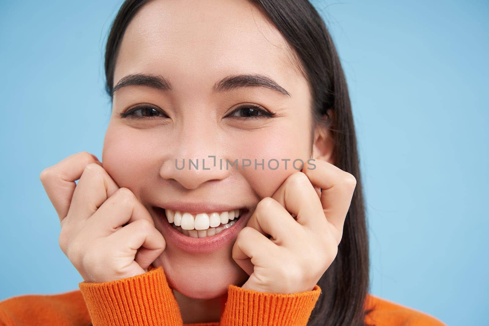 Close up portrait of beautiful japanese brunette girl, smiling and looking lovely at camera, gazing with care and tenderness, blue background.