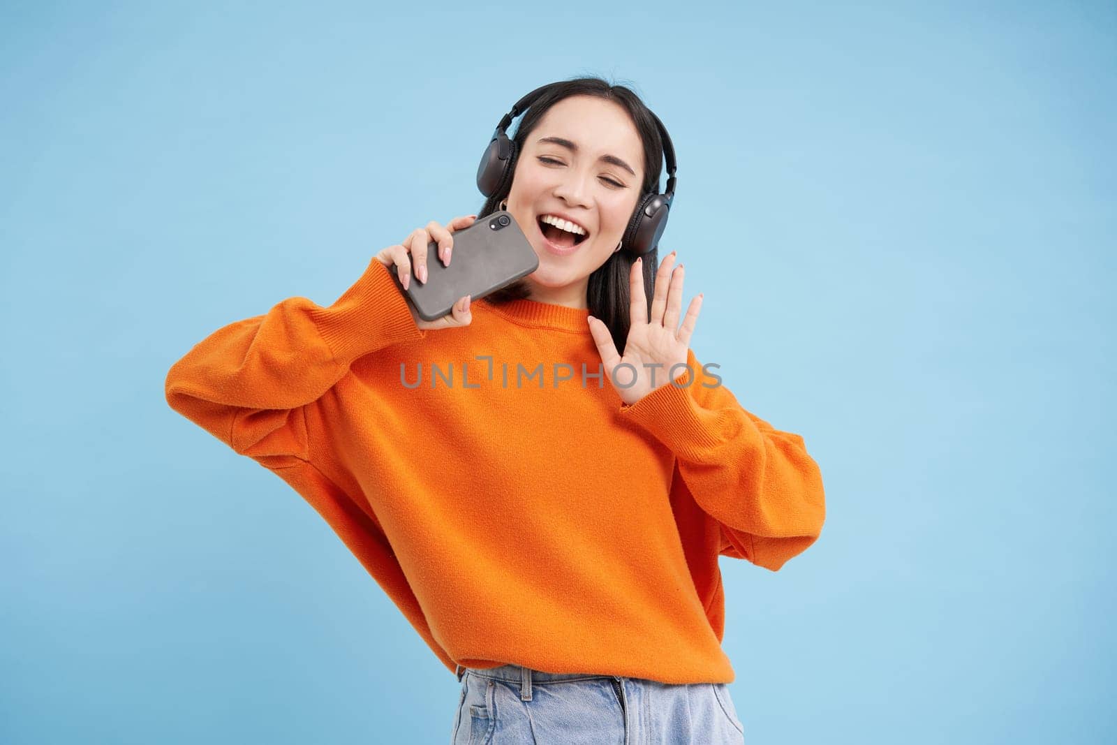 Beautiful korean girl singing in smartphone mic, listens music in headphones, sings karaoke on mobile app, stands over blue background.