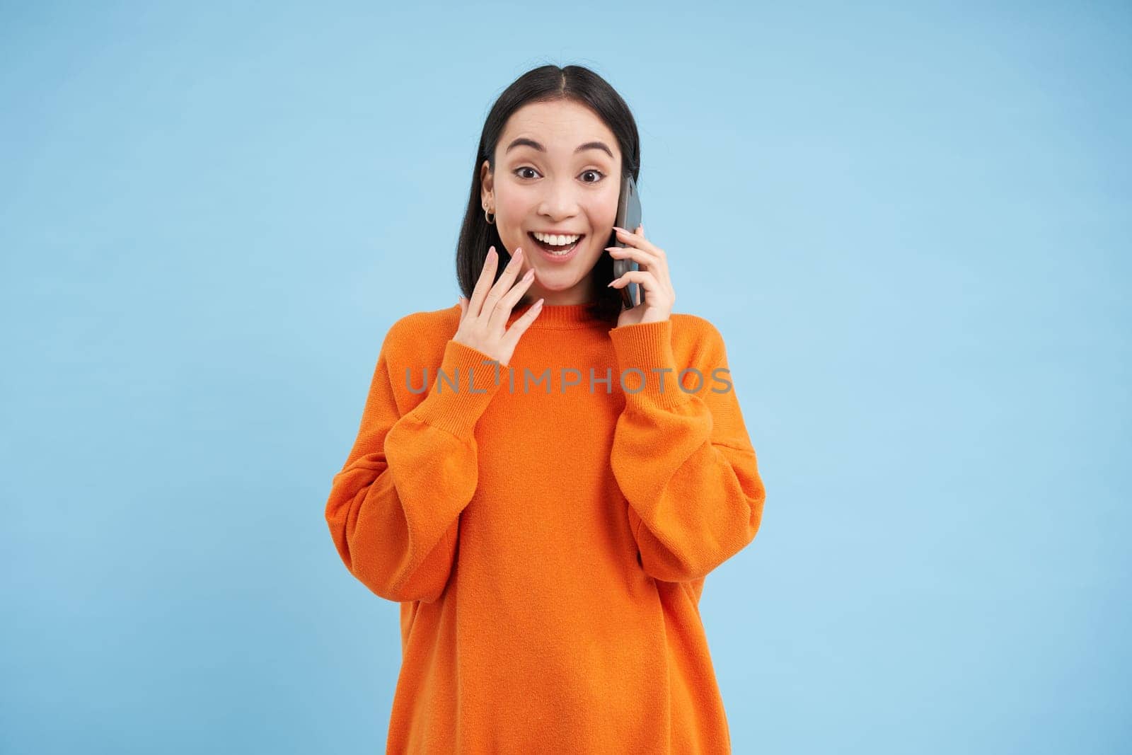 Surprised korean woman answers phone call and reacts to great news, celebrating, standing over blue studio background.