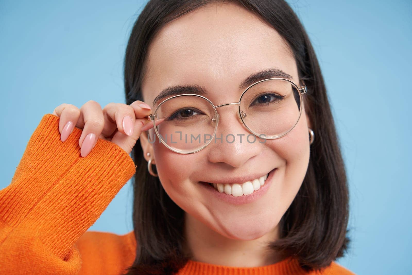 Portrait of smiling beautiful woman in glasses, looks happy at camera, standing over blue studio background. Opticians and eyewear concept