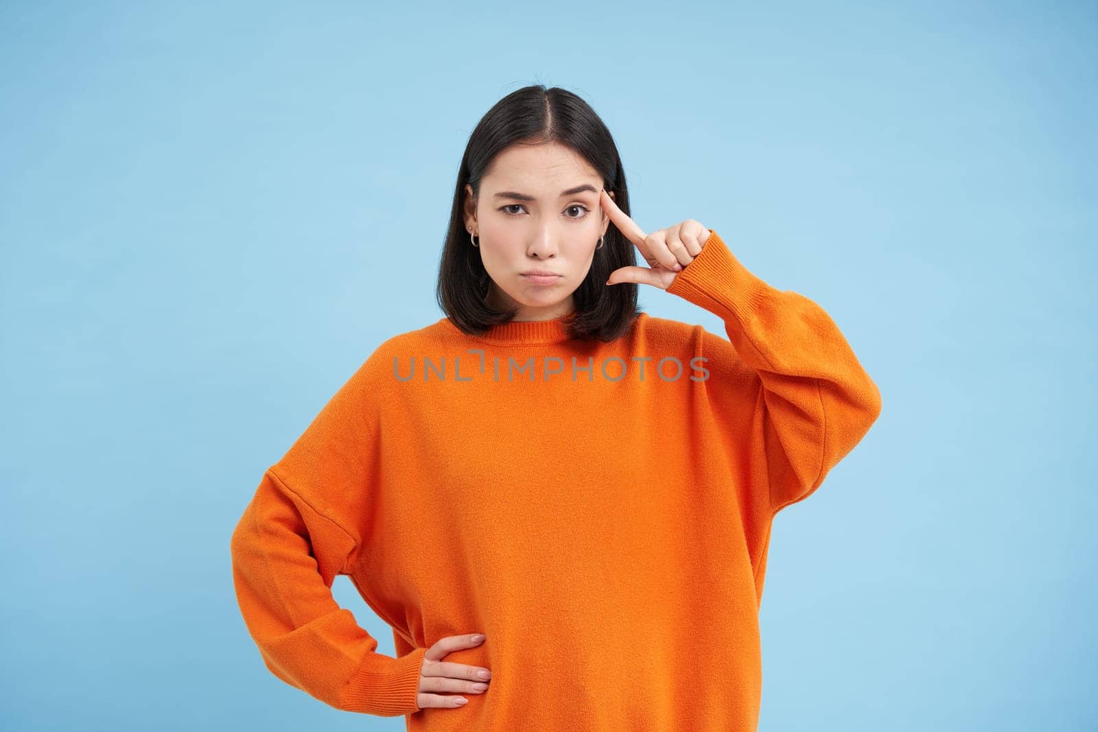 Are you crazy. Young skeptical asian woman holds finger near head, disappointed by someones behaviour, stands over blue background.