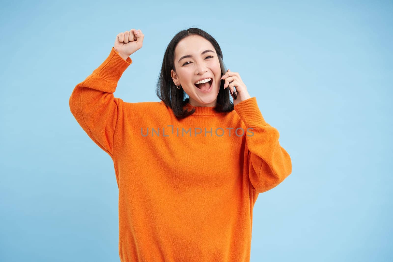 Happy asian girl receives excellent news over the phone, holds mobile and does fist pump, celebrates victory, achievement, blue studio background.