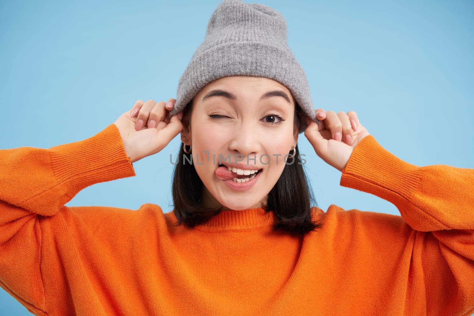 Close up portrait of Asian female model puts on hat and winks at you, looking happy, going outside, standing over blue background.