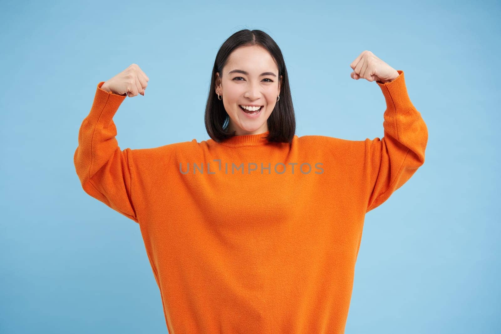 Strong and healthy people. Young asian woman shows her muscles, flexing biceps and looks confident, smiling pleased, in great shape, blue background.