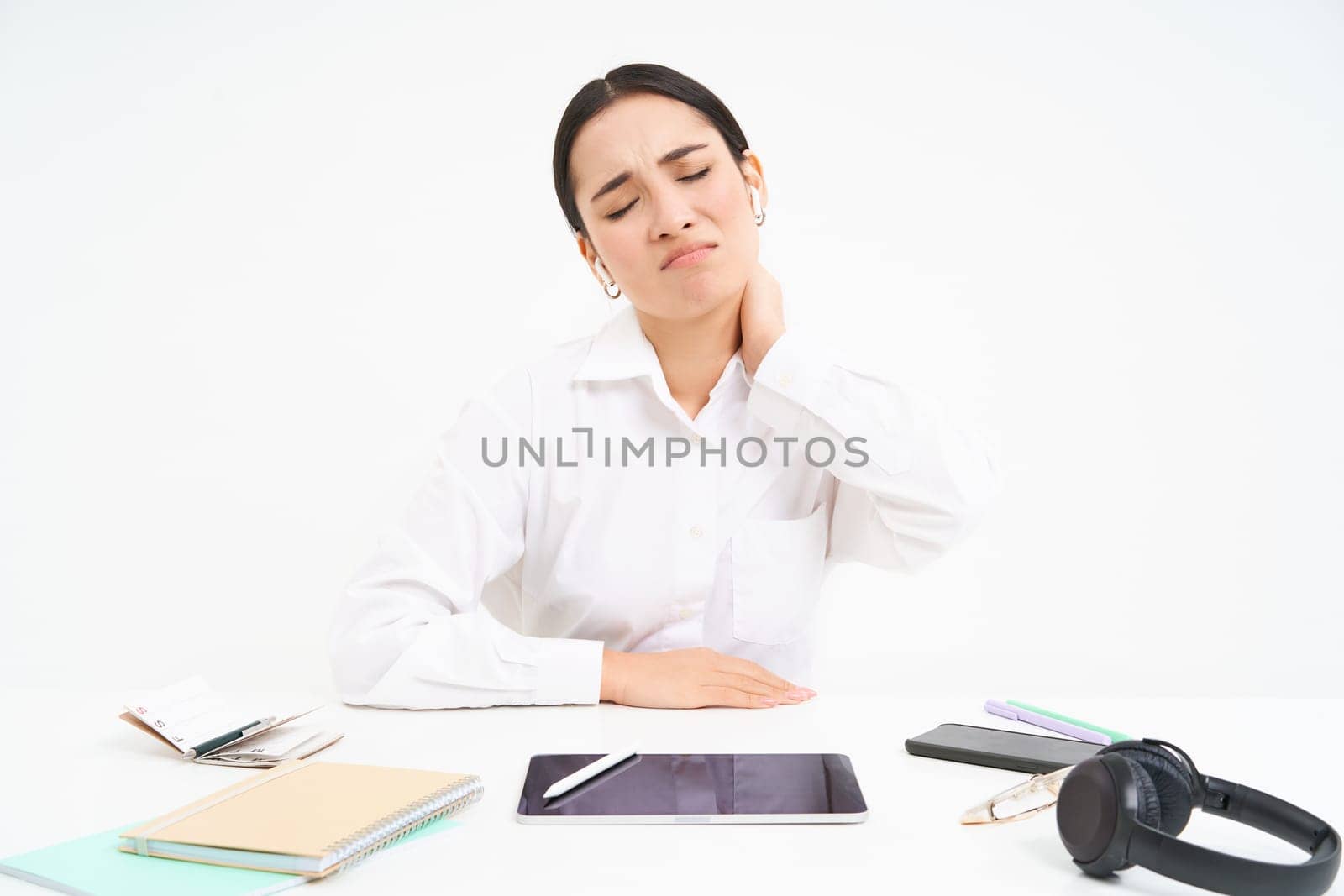 Tired asian woman, office lady sits behind her desk, has back pain, massaging her shoulder, tired after work, white background.